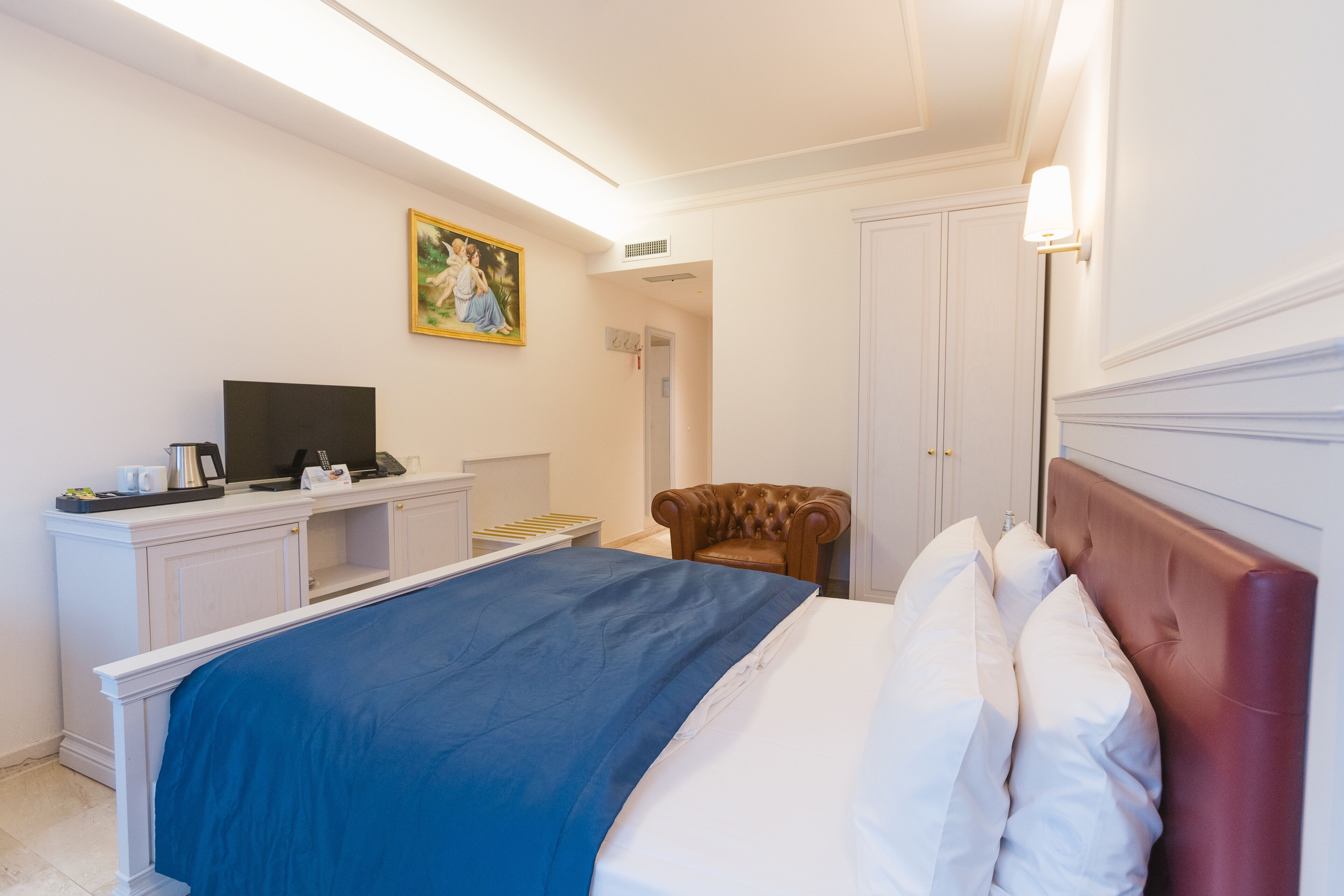 A hotel room with a bed, a desk with a computer, a chair, and a painting on the wall.