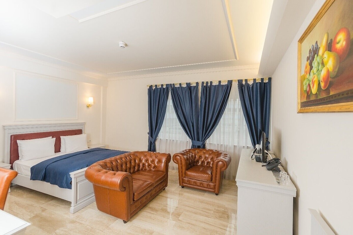 A hotel room with a bed, two leather armchairs, a painting, and a desk with a lamp.