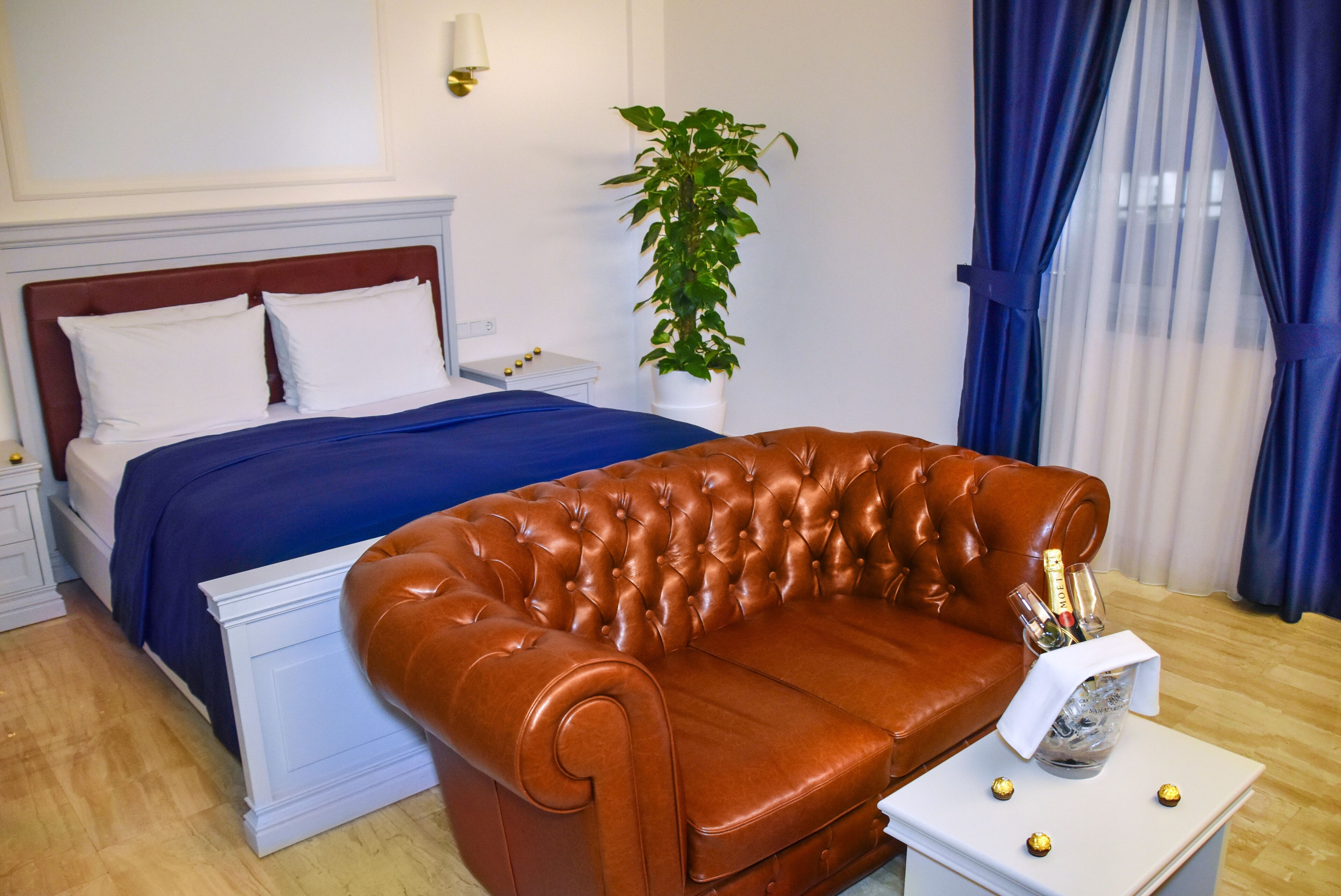 A bedroom with a bed, a brown leather sofa, a bedside table with a bottle of champagne, and a potted plant.