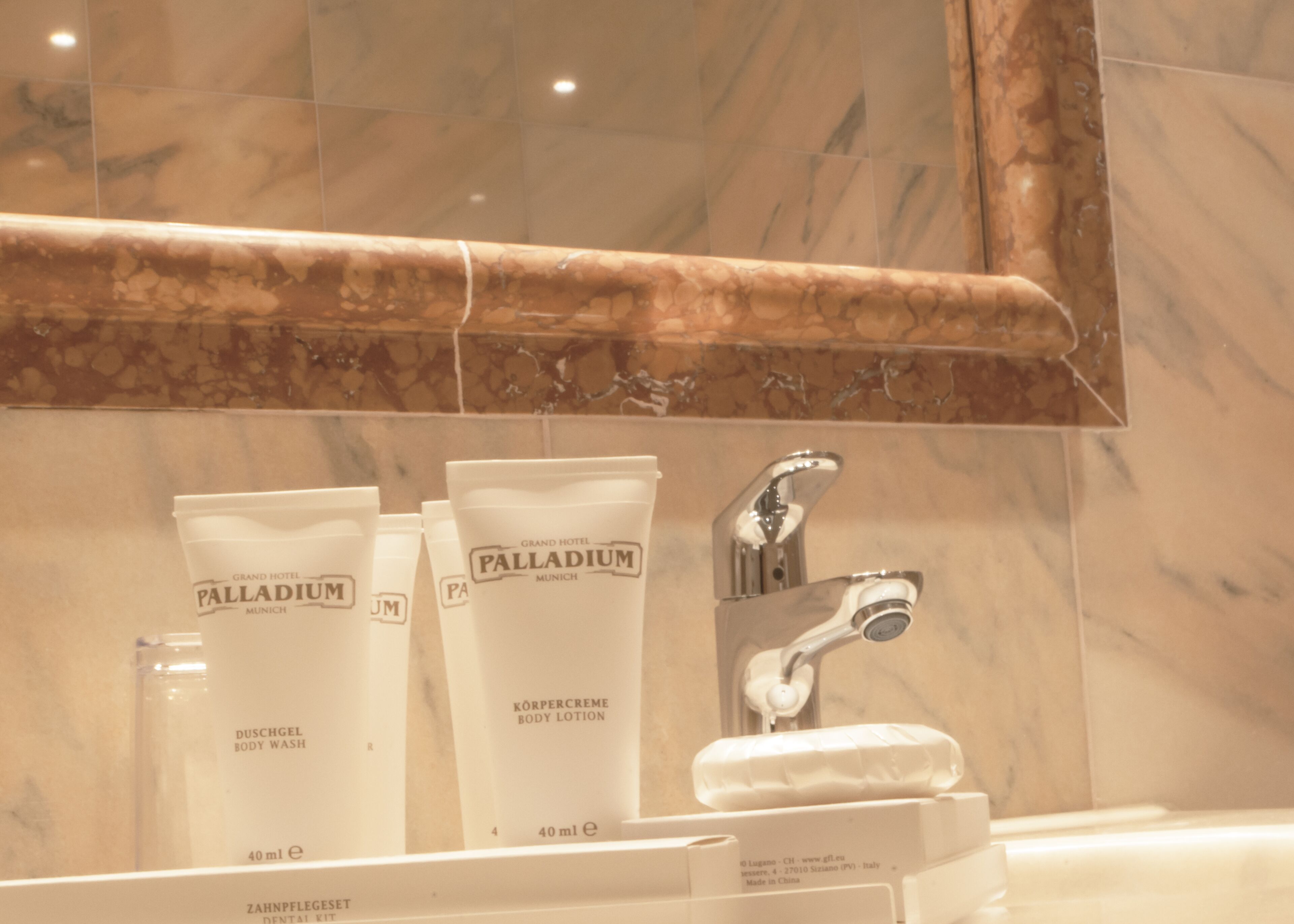 A bathroom with a marble sink, a mirror, and three bottles of Palladium body lotion.