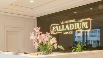 Un comptoir d’accueil avec une enseigne indiquant « Grand Hôtel Palladium Munich » et un vase de fleurs.