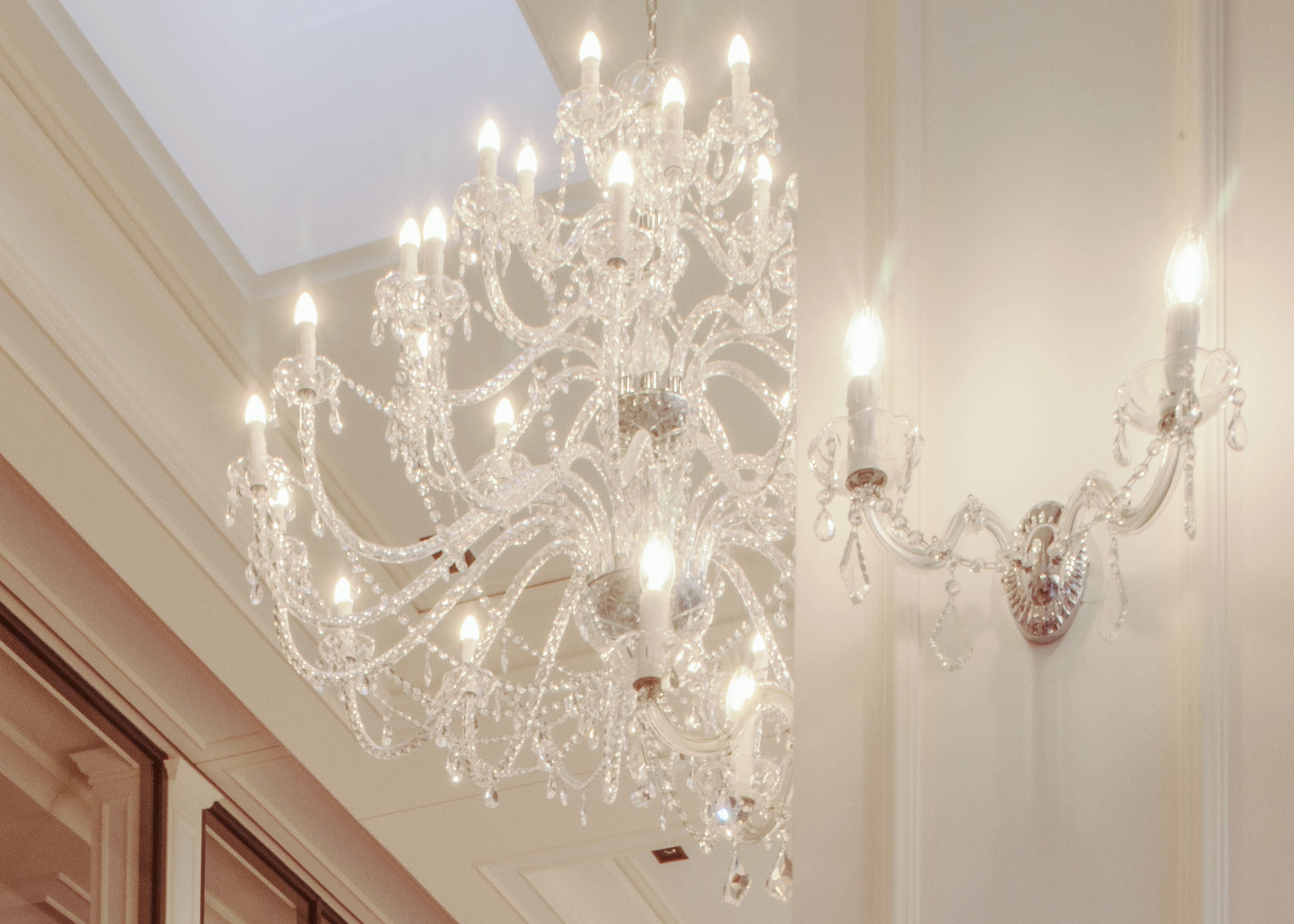 A grand chandelier with multiple lights and crystal decorations, hanging from a high ceiling in a spacious room with elegant architecture.
