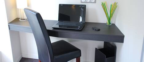 Escritorio moderno con una computadora portátil, una silla, una lámpara y un florero con ramas verdes y amarillas.