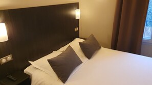 A hotel room with a large bed, two pillows, a nightstand with a remote control, and a wall-mounted light.