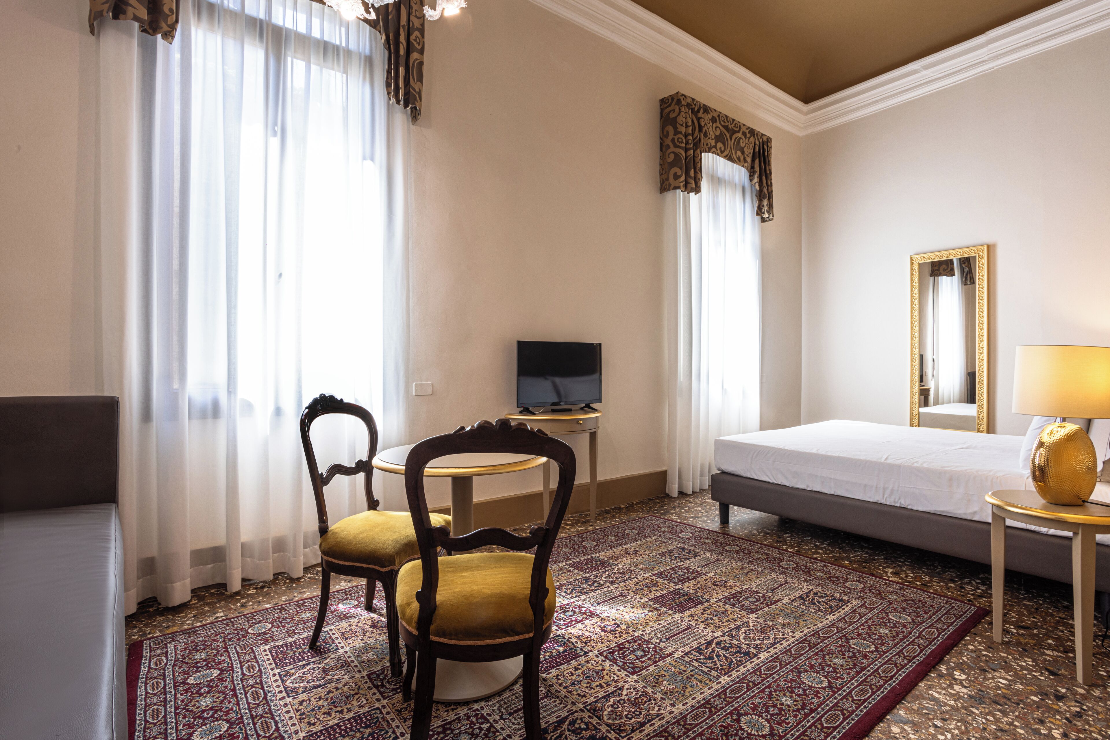Une chambre d’hôtel avec un lit, un téléviseur posé sur un meuble, une chaise, une table avec une lampe et un grand miroir.