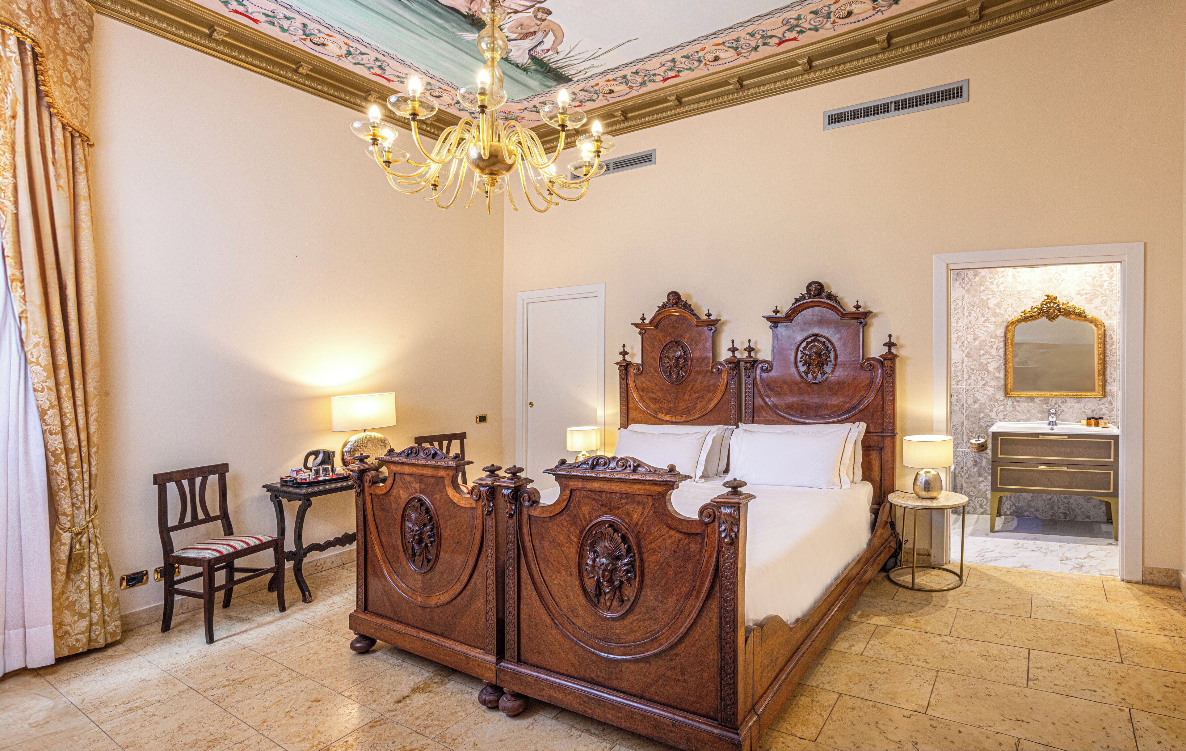 Ruime slaapkamer met een groot bed, een kroonluchter, een kaptafel met spiegel, en een apart zithoekje met een stoel en tafel.