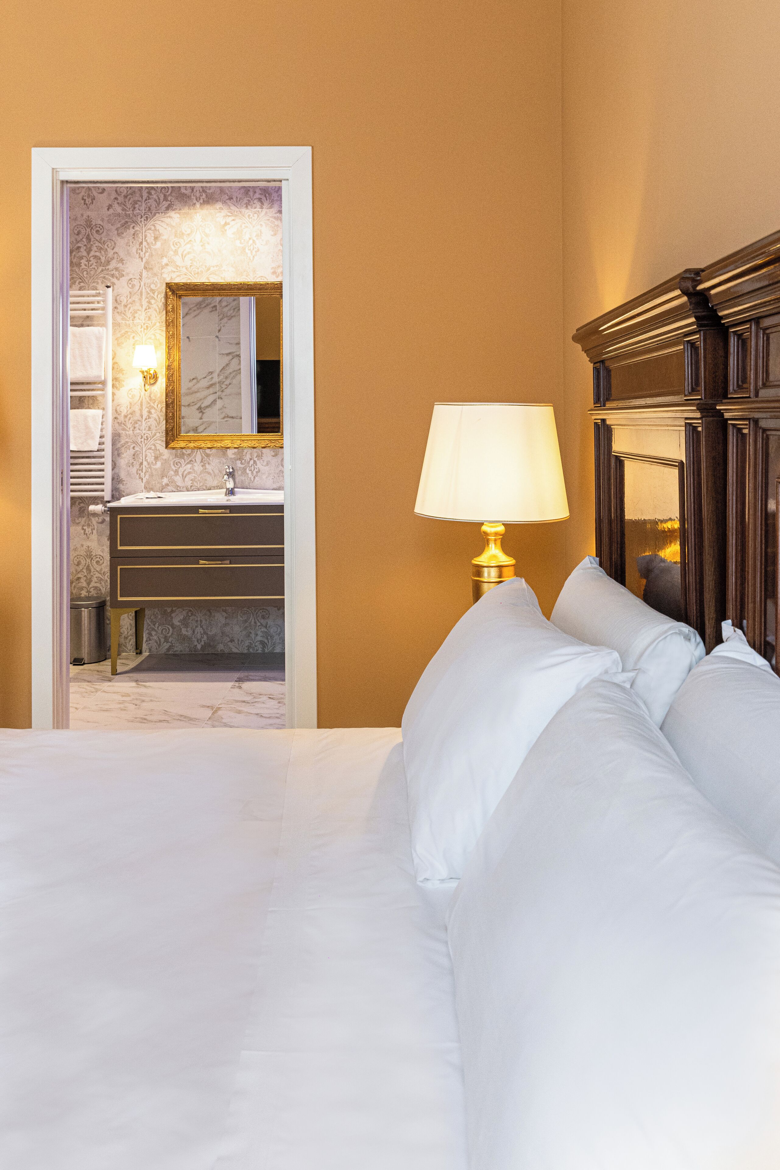 A hotel room with a bed, a nightstand with a lamp, a bathroom with a sink and mirror, and a door leading to another room.