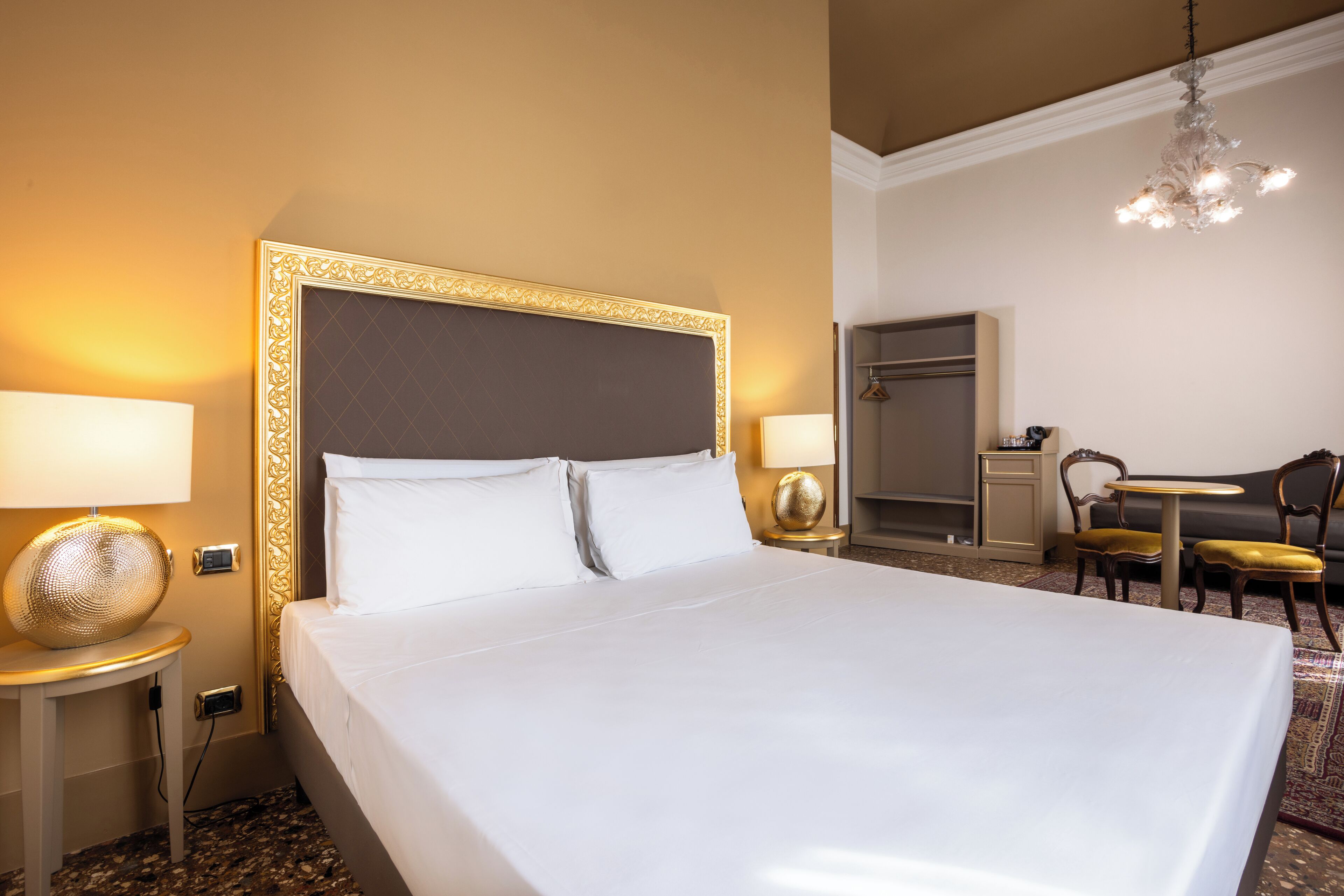 Habitación de hotel con una cama grande, dos lámparas de noche, una araña, un escritorio con una silla y una mesita con otra silla.