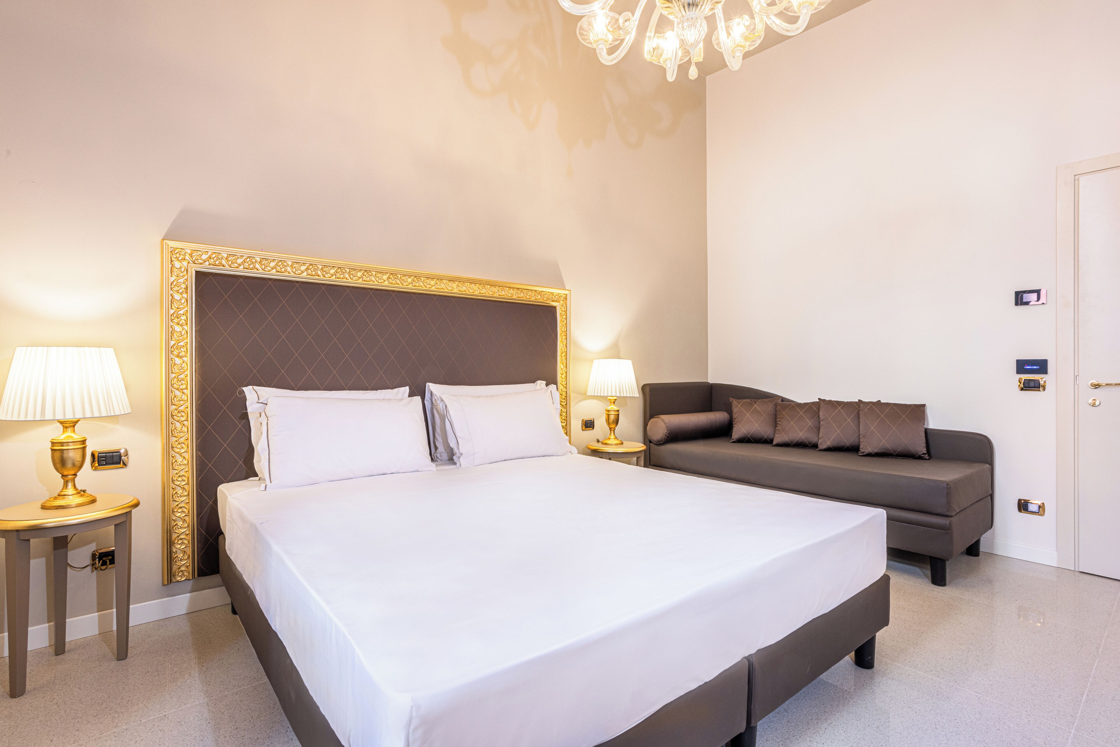 A hotel room with a large bed, a sofa, two bedside tables with lamps, and a chandelier.