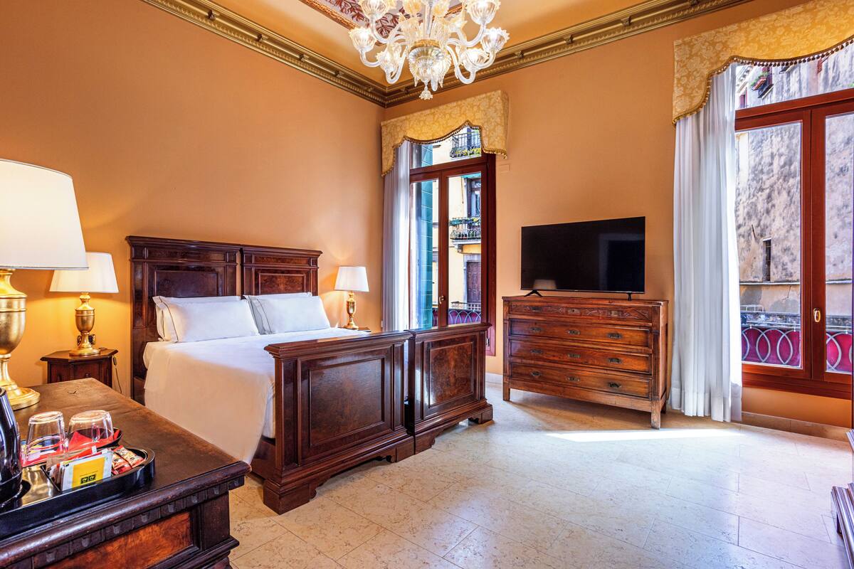 Ein Hotelzimmer mit einem großen Bett, einem Holzkapstativ, einem Fernseher und einem Balkon mit Blick.