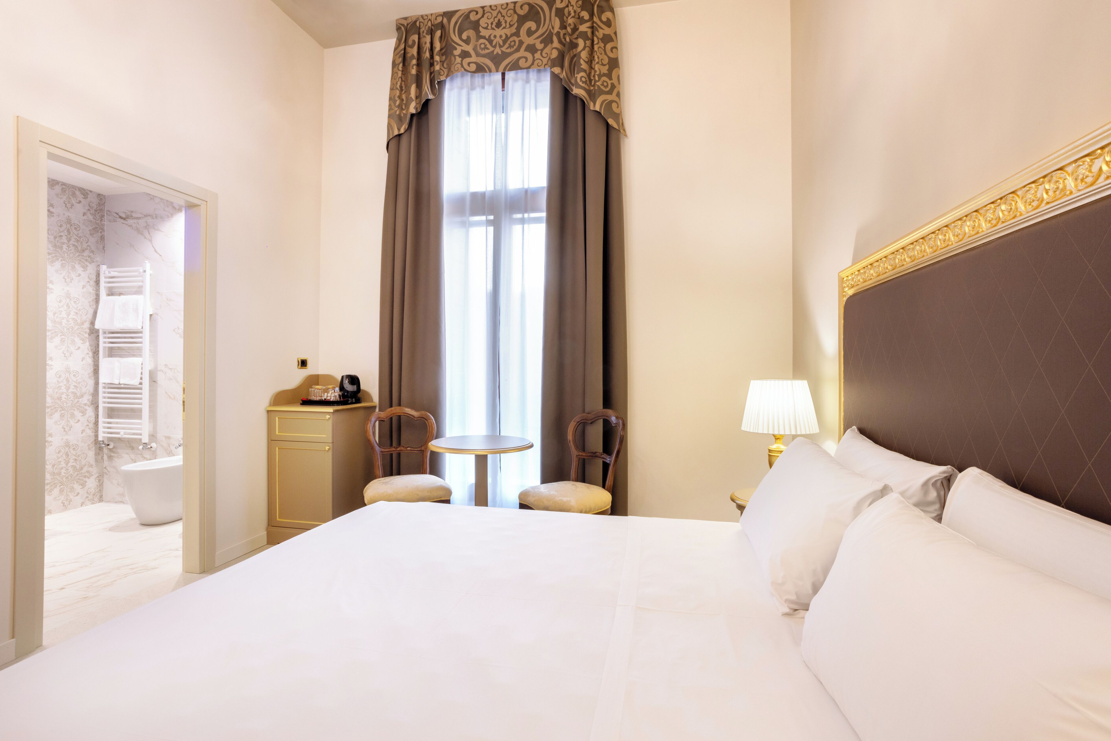 A hotel room with a large bed, two chairs, a small table, a lamp, and a view of a bathroom with a bathtub and towel rack.
