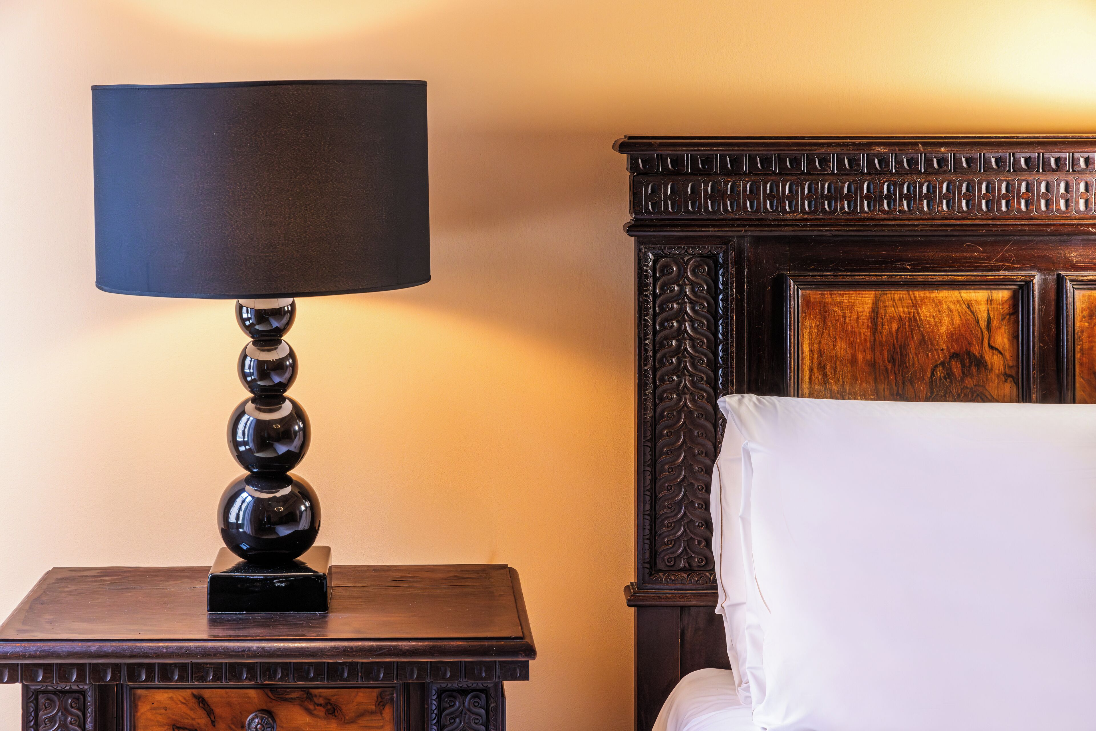 A wooden nightstand with a black lamp and a white pillow on a bed with a carved headboard.