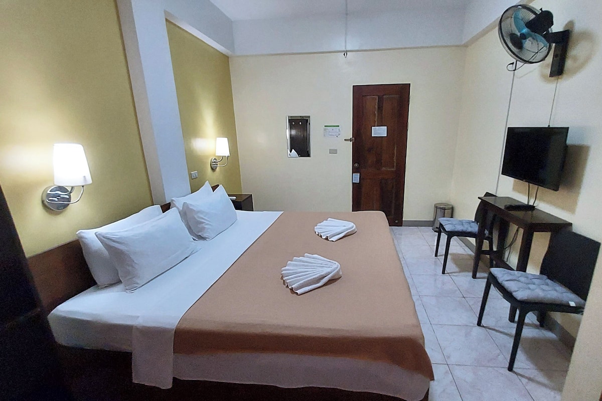 A hotel room with a bed, a desk, a chair, a television, and a wall-mounted fan.