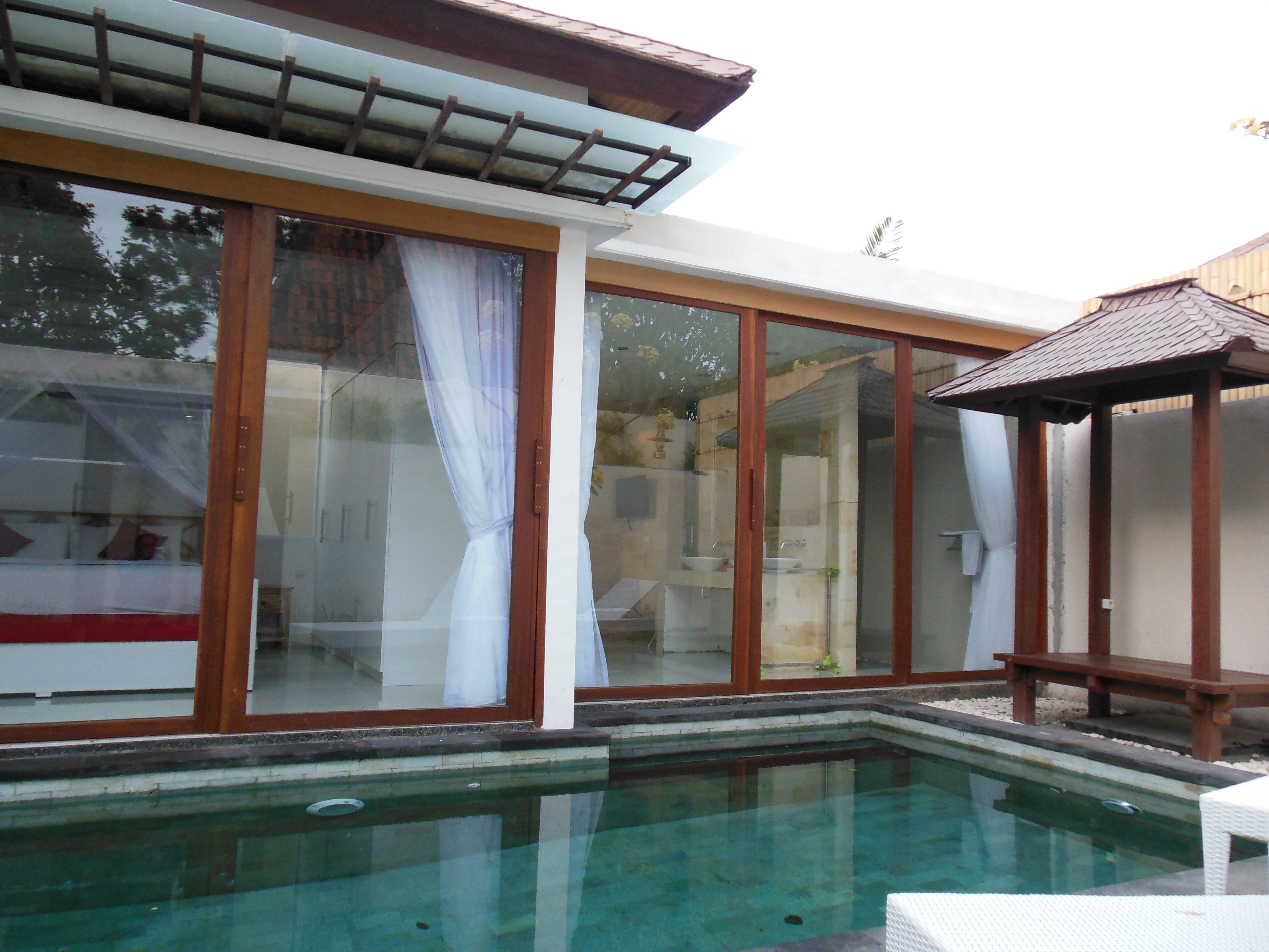 Una piscina con cerramiento de vidrio, una pérgola de madera y vistas a un edificio con puertas y cortinas de vidrio.