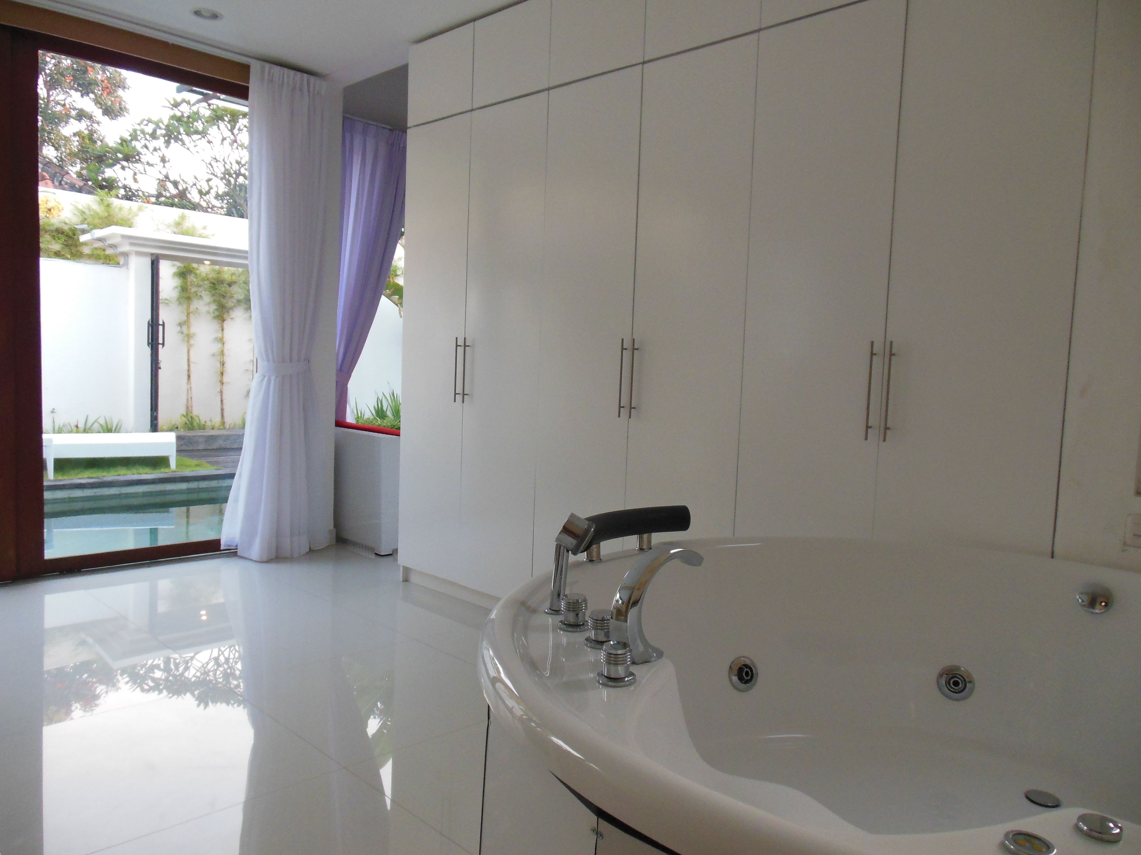 A modern bathroom with a large bathtub, white cabinets, and a view of a pool and greenery outside.