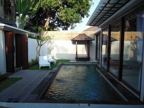 Un área de piscina moderna al aire libre con terraza de madera, dos sillones blancos, una mesita de madera y una pérgola.