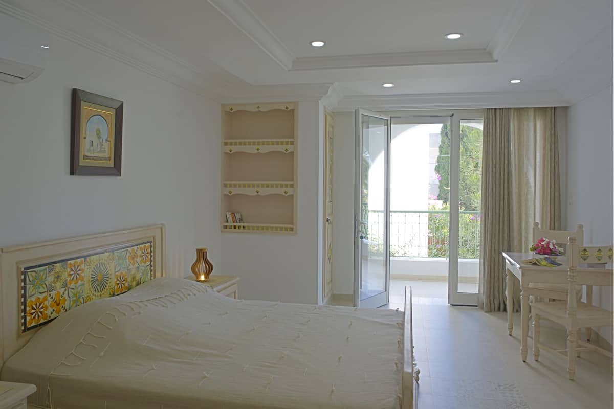 A bedroom with a large bed, a bedside table, a chair, a small table, a picture on the wall, and a balcony with a view.