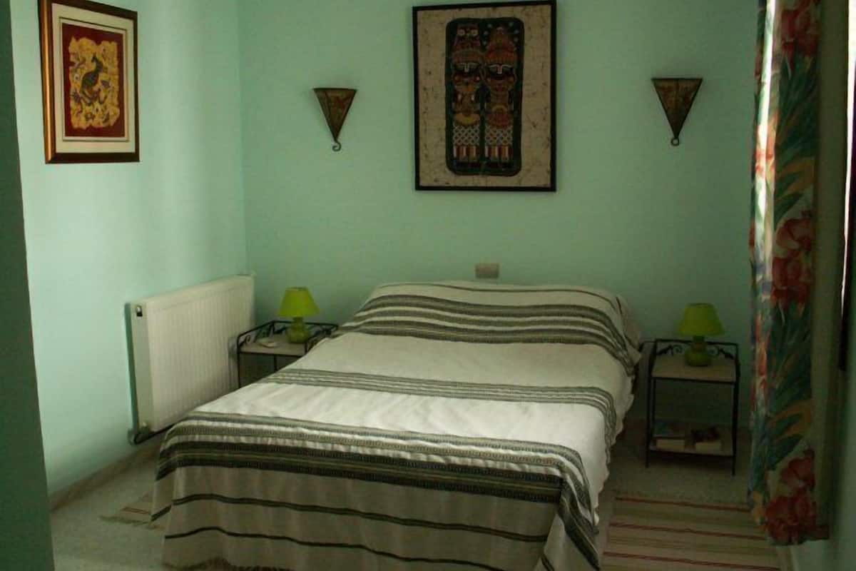 A bedroom with a bed, two bedside tables with lamps, a radiator, and two wall-mounted wall art pieces.