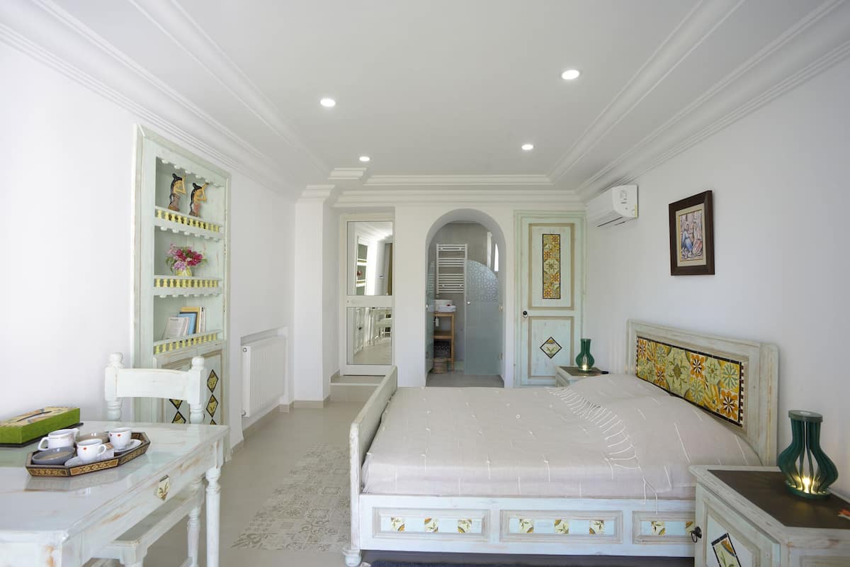 A bedroom with a bed, a dresser, a vanity, and a shelf with decorative items.