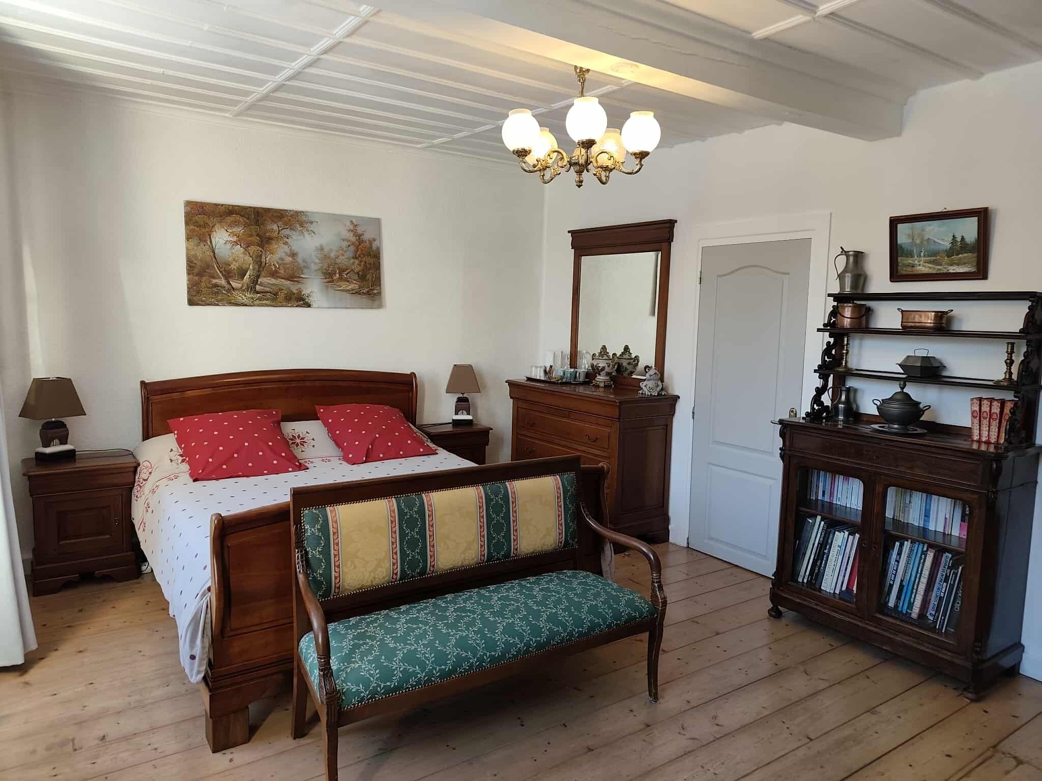 Un dormitorio con una cama de madera, un banco, un aparador con libros, un espejo y una araña.