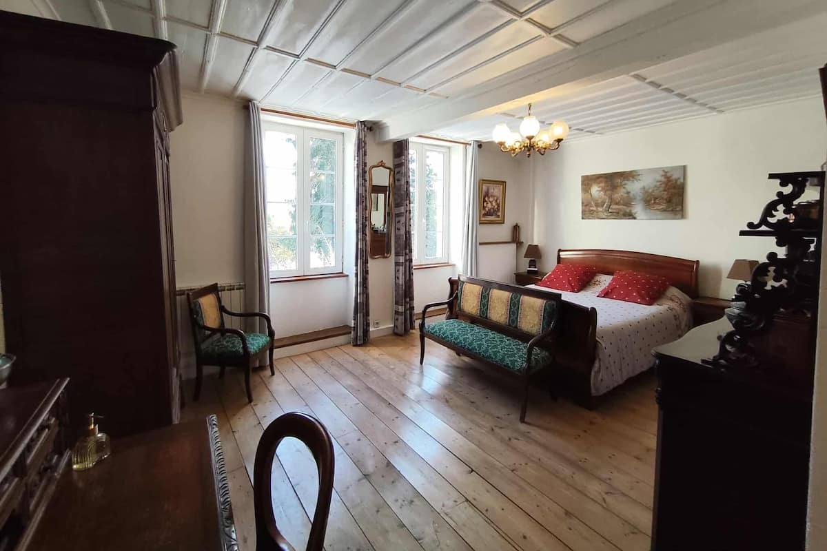 Une pièce avec un plancher en bois, un lit recouvert d’une courtepointe rouge et blanche, une chaise en bois, un bureau en bois et un lustre.
