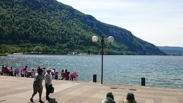 A lakeside promenade with people walking and sitting on benches, overlooking a calm lake and a forested hillside.