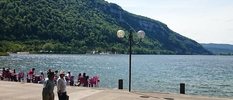 Un paseo marítimo junto al lago con gente caminando y sentada en bancos, con vistas a un lago tranquilo y una ladera boscosa.
