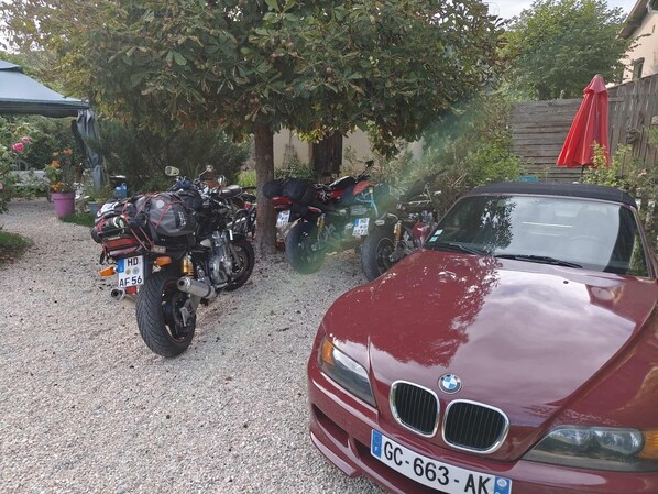 A maroon BMW car parked on a gravel driveway with motorcycles and a red umbrella in the background.