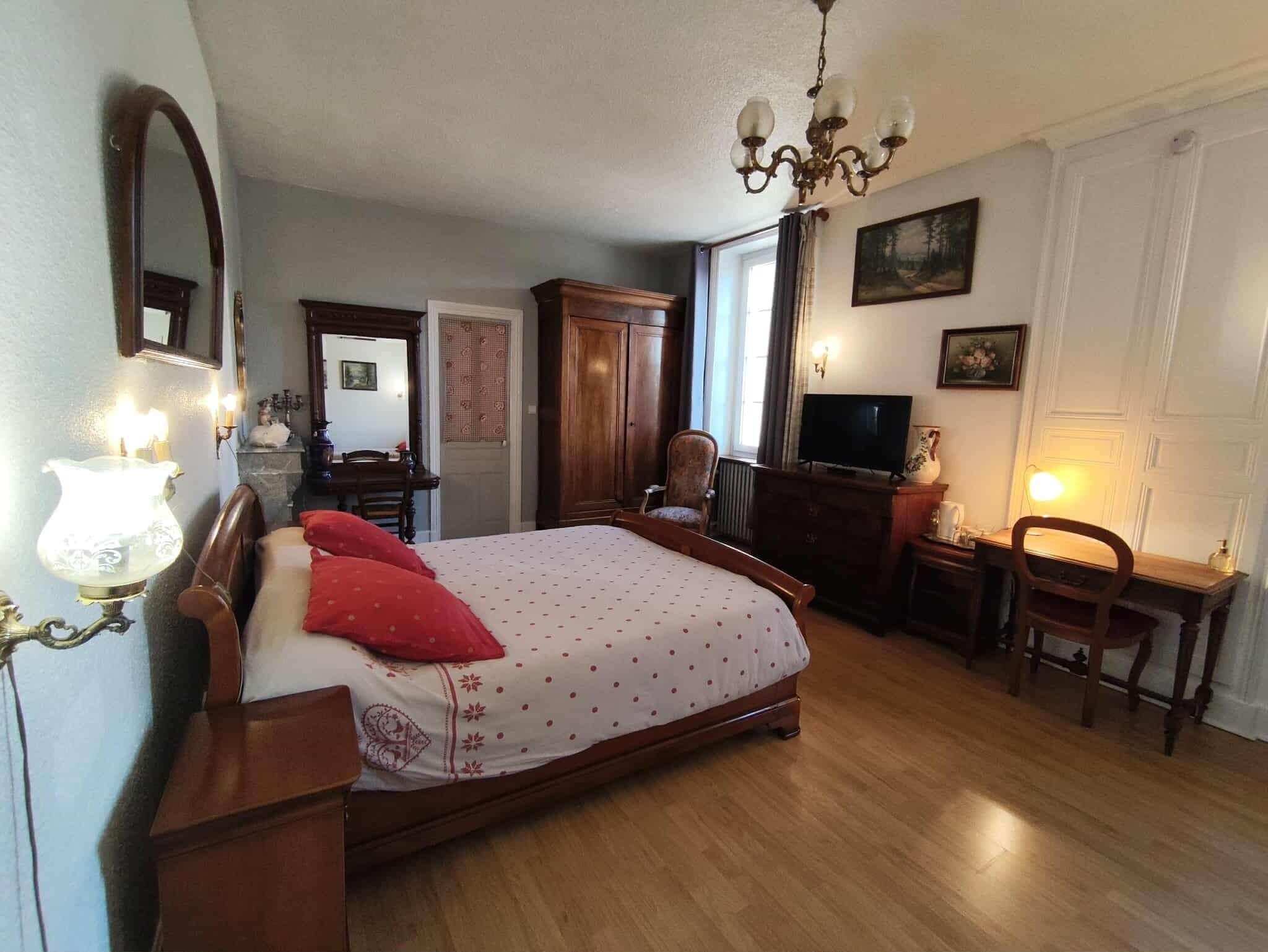 A bedroom with a wooden bed, a desk, a chair, a television, and a chandelier.