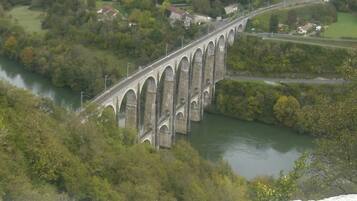 Eine steinerne Brücke überspannt einen Fluss, umgeben von Grünpflanzen und Häusern.
