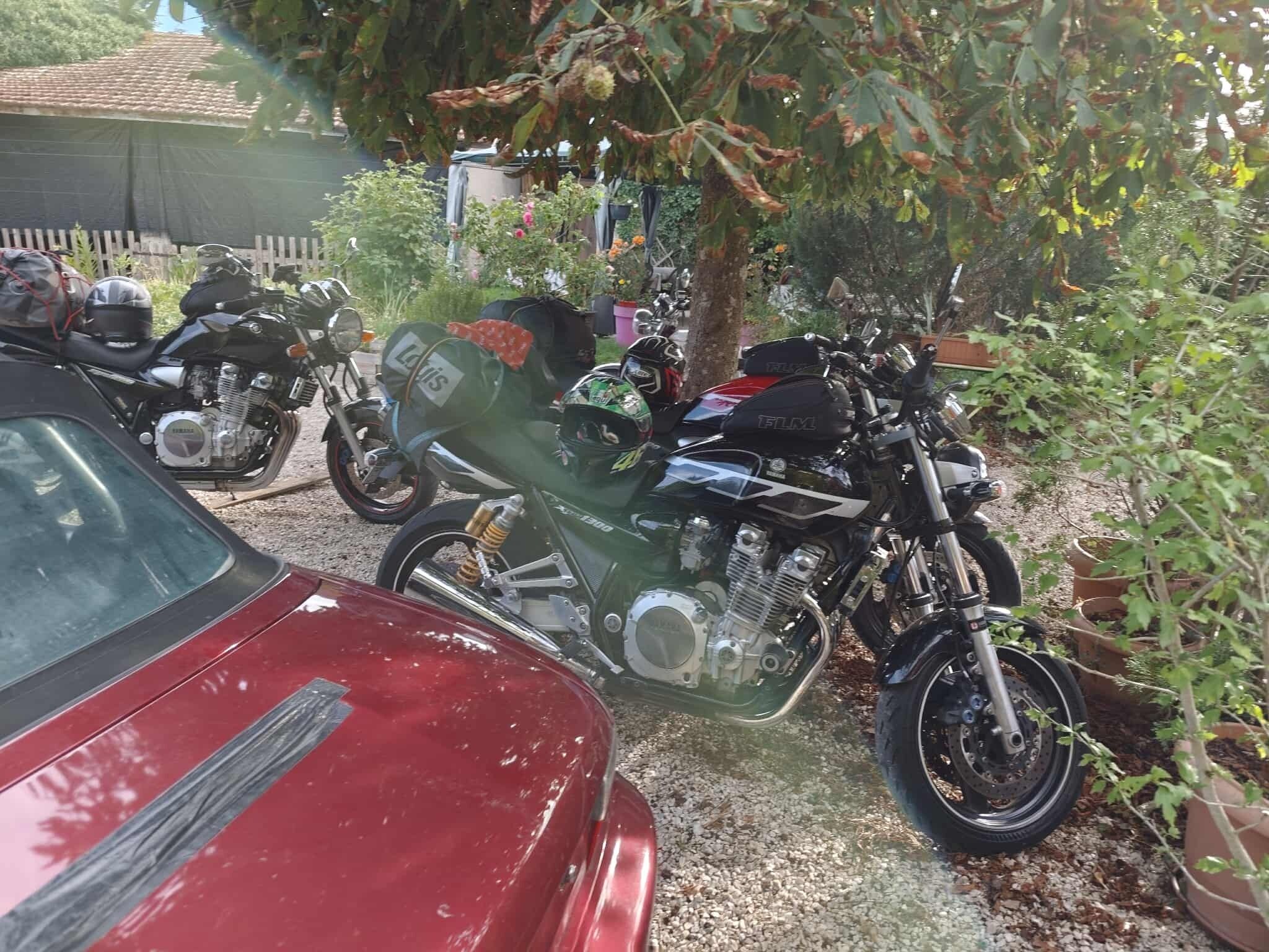 A red car parked next to motorcycles in a garden area with trees and a fence in the background.