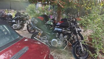 A red car parked next to motorcycles in a garden area with trees and a fence in the background.