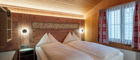 A wooden cabin room with two beds, a nightstand, a wall-mounted heater, and a window with curtains.