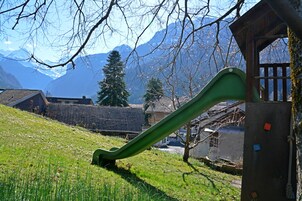 Un terrain de jeux avec un toboggan, entouré d’une pelouse, de bâtiments et, au loin, de montagnes.