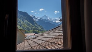 Blick durch ein Fenster auf ein Gebäude mit traditionellen, holzverkleideten Satteldächern, eine Berglandschaft und einen klaren Himmel.