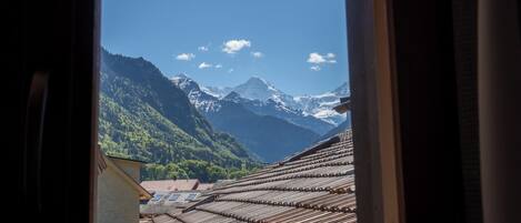 Vista a través de una ventana que muestra un edificio con techos de madera inclinados, un paisaje montañoso y cielo despejado.