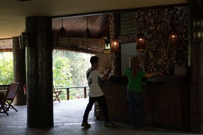 A reception area with a stone wall and wooden pillars, a counter, and a view of greenery outside. - Samboja Lodge (Balikpapan)