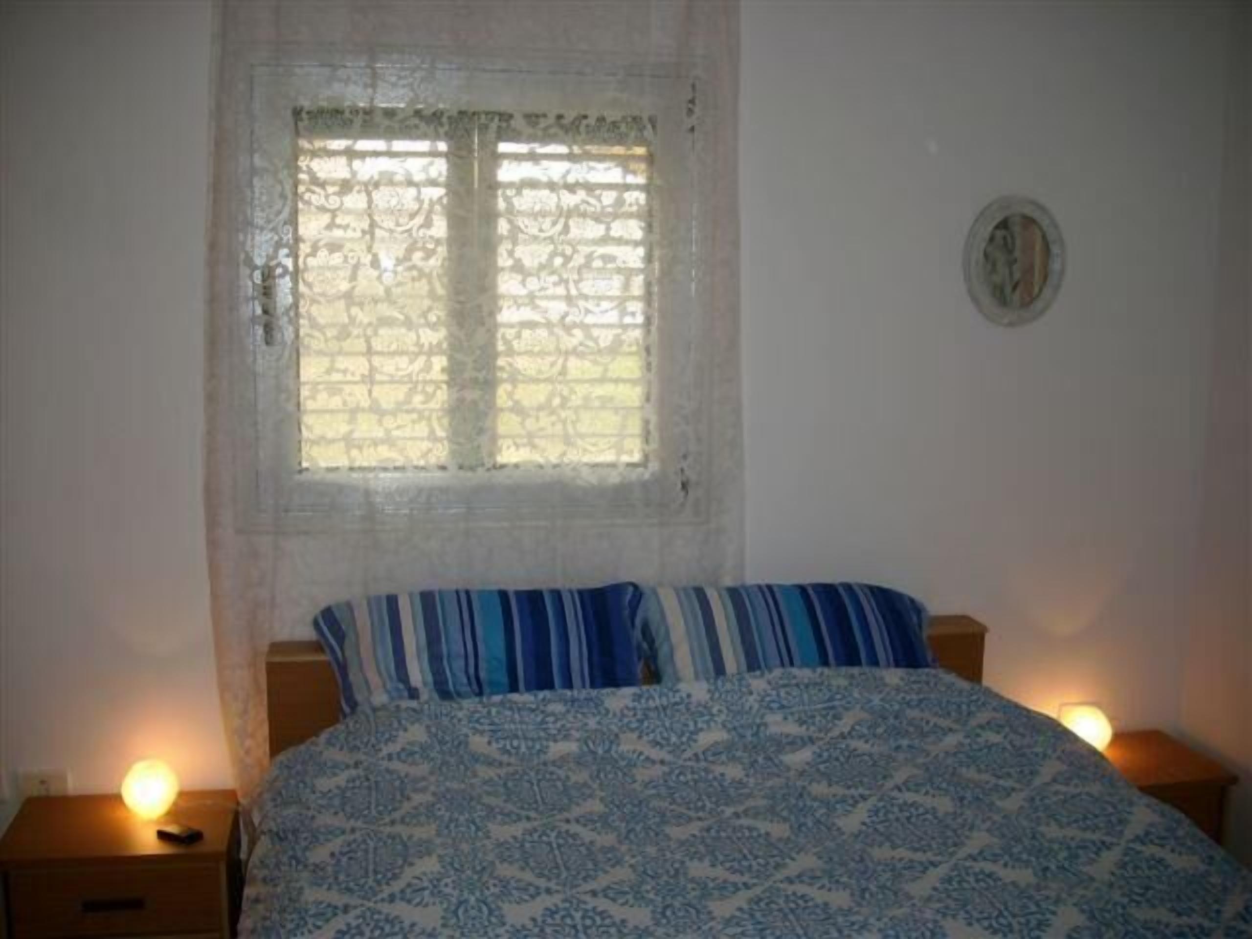 A bedroom with a bed, a window with lace curtains, two bedside tables with lamps, and a wall clock.
