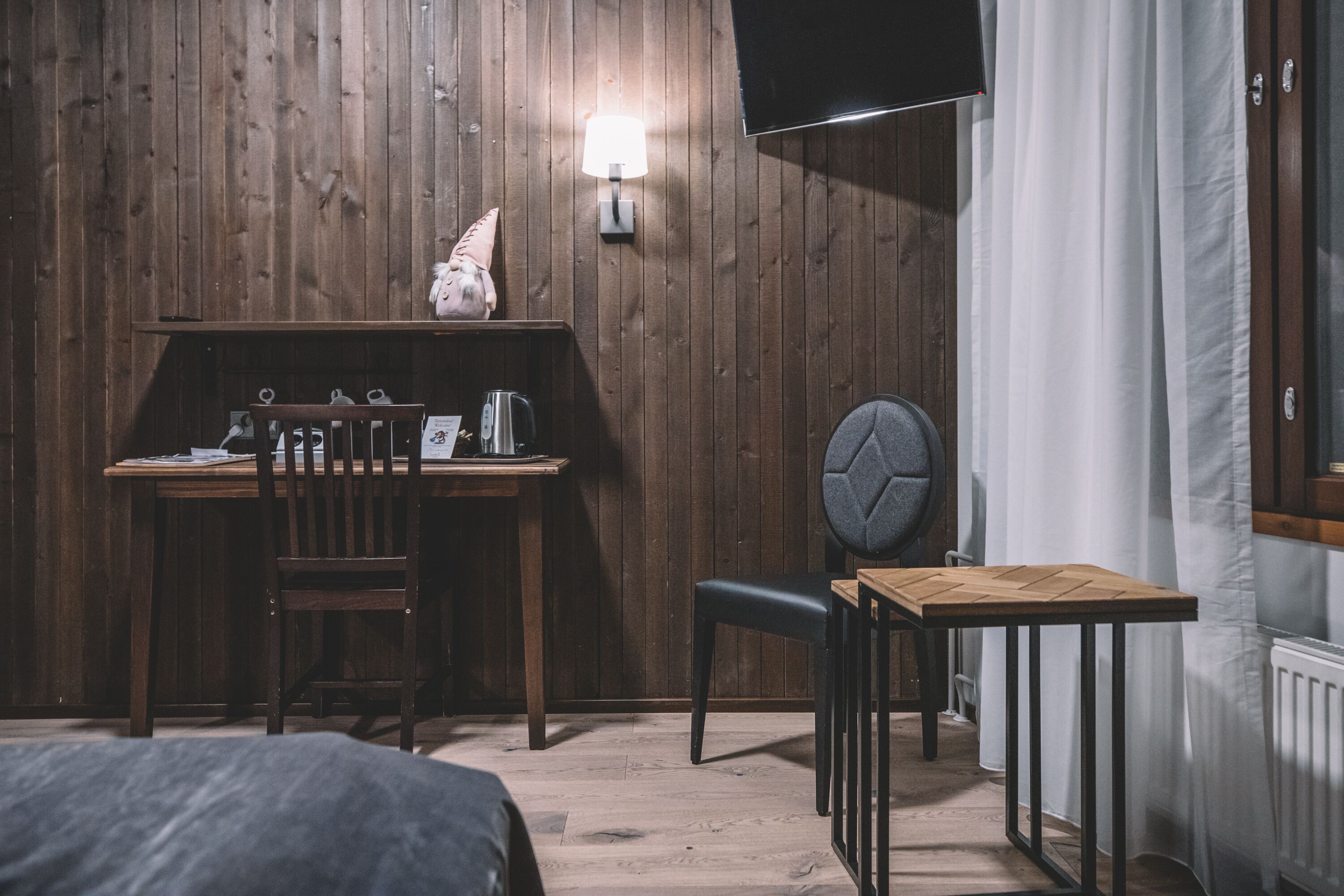 A room with a wooden wall, a desk with a chair, a small table with a chair, a lamp, and a bed.