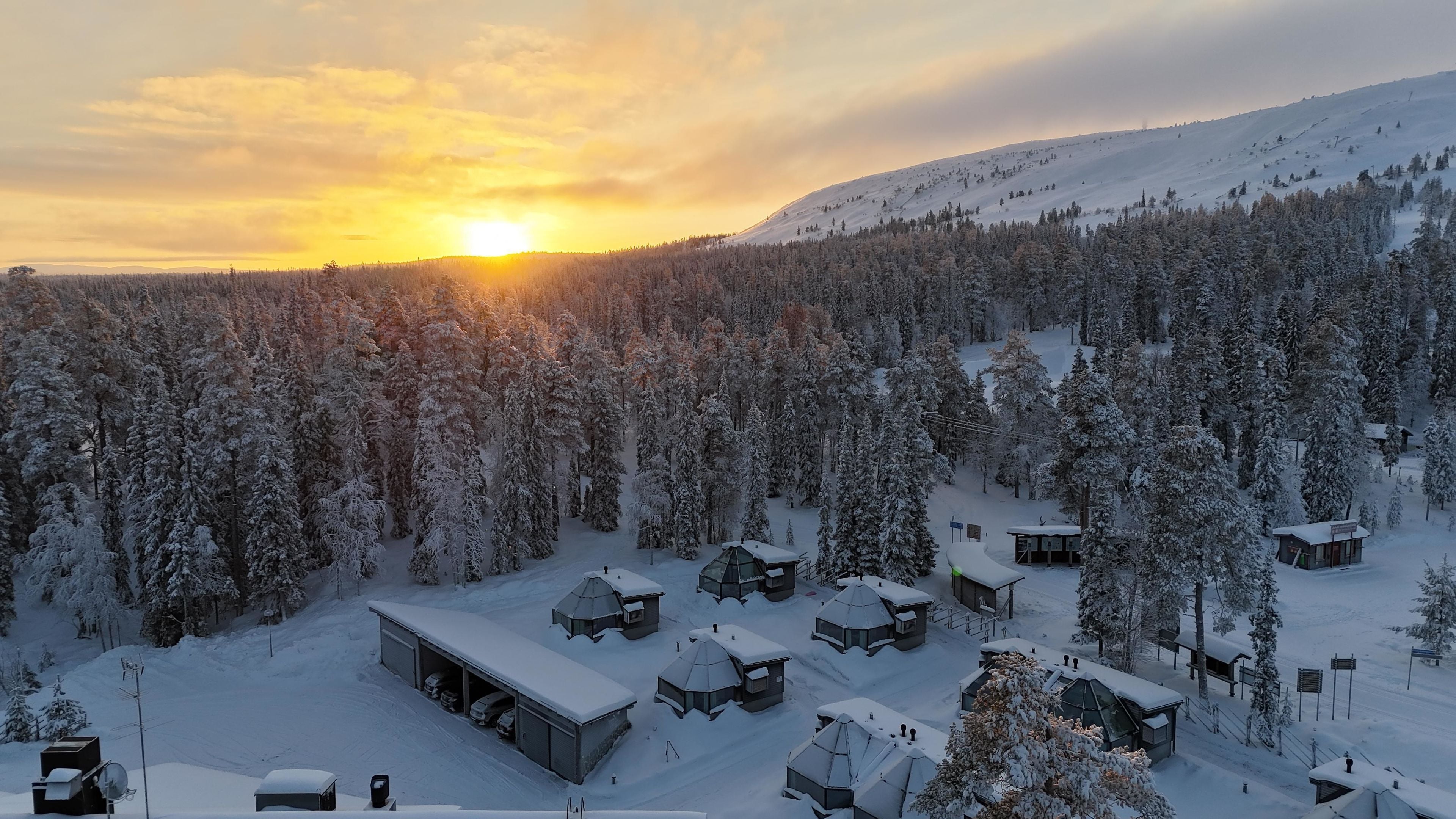 Lumipeitteinen maisema, jossa on useita jurttatelttaa ja suuri rakennus, todennäköisesti majoitusrakennus, ja taustalla näkyy metsäinen kukkula.