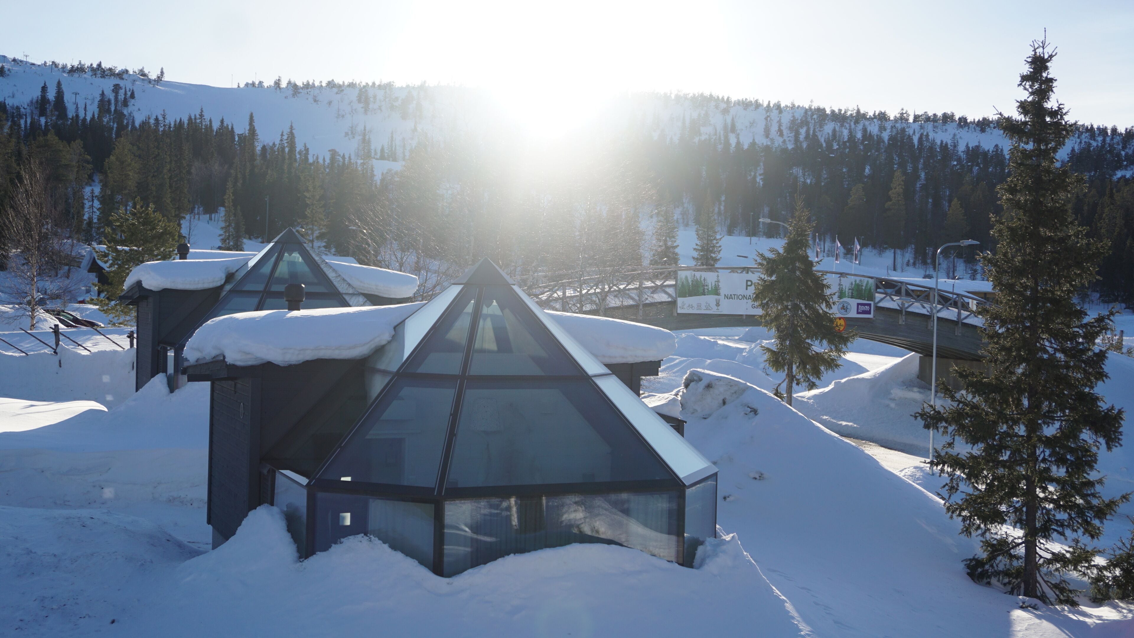 A modern building with a glass roof amidst a snowy landscape, surrounded by trees and a clear sky.