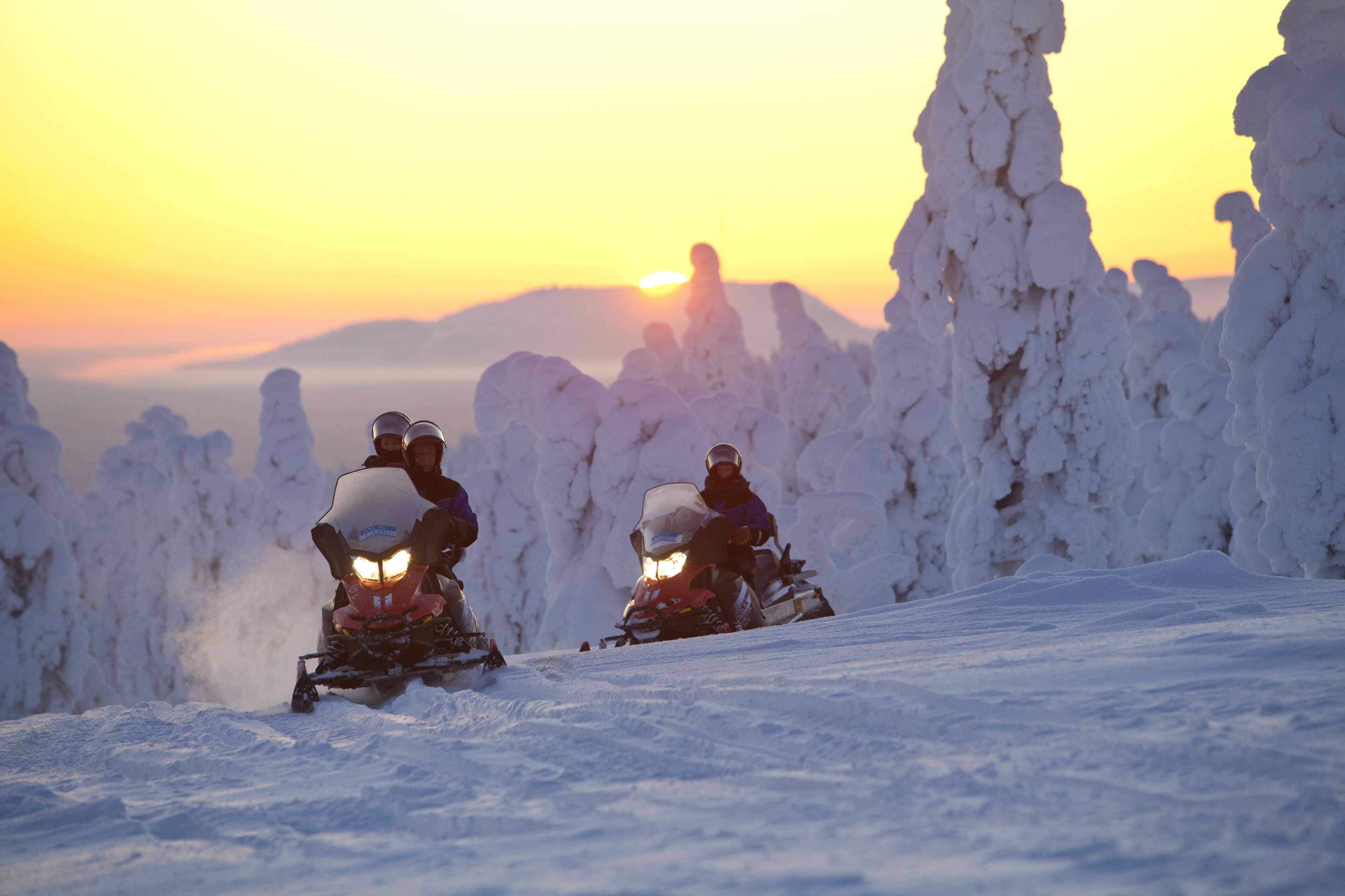 Two snowmobiles on a snowy slope with a sunset in the background.