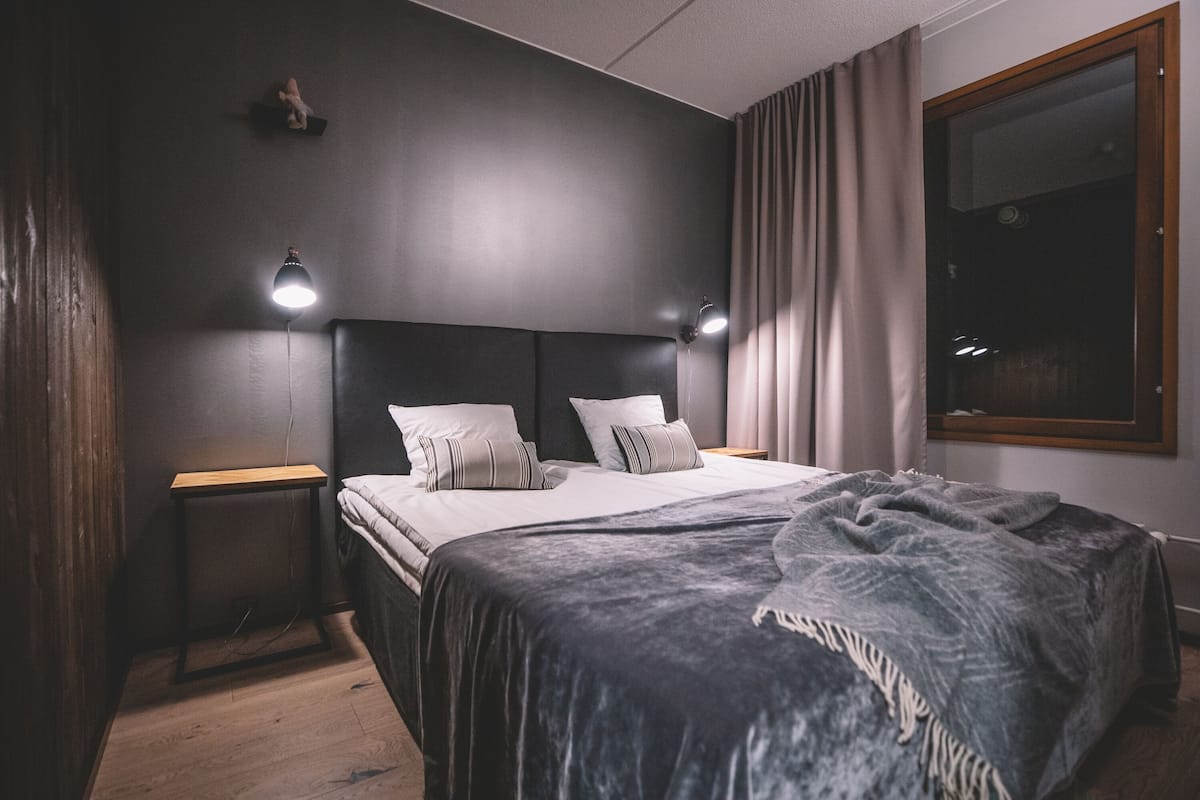 Un dormitorio con cama, mesitas de noche, lámpara, ventana con cortinas y un pequeño estante con una figurita.
