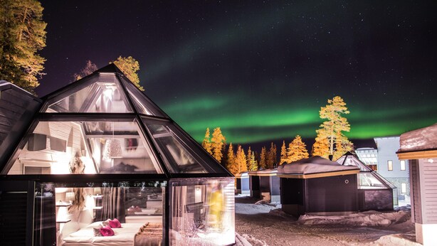 Lasikatolla varustettu mökki, jonka sisätilat ovat näkyvissä, ja jonka ympärillä on lumipeitteinen maa ja puita. Yllä on tähtikirkas yötaivas ja revontulia.