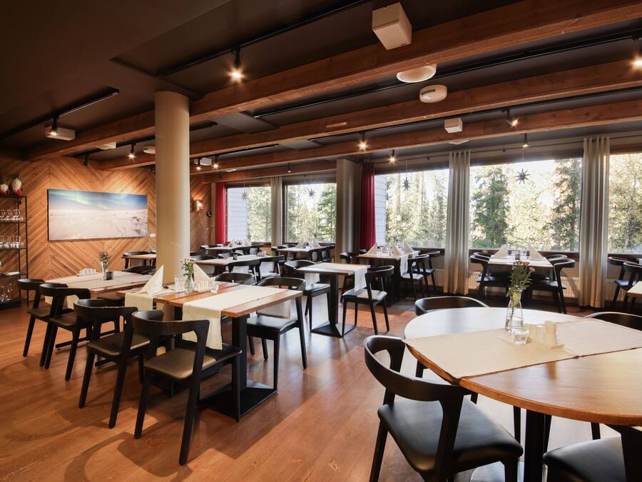 Een ruime eetzaal met houten vloeren en tafels, grote ramen met gordijnen, en een visgraatmotief aan de wand.