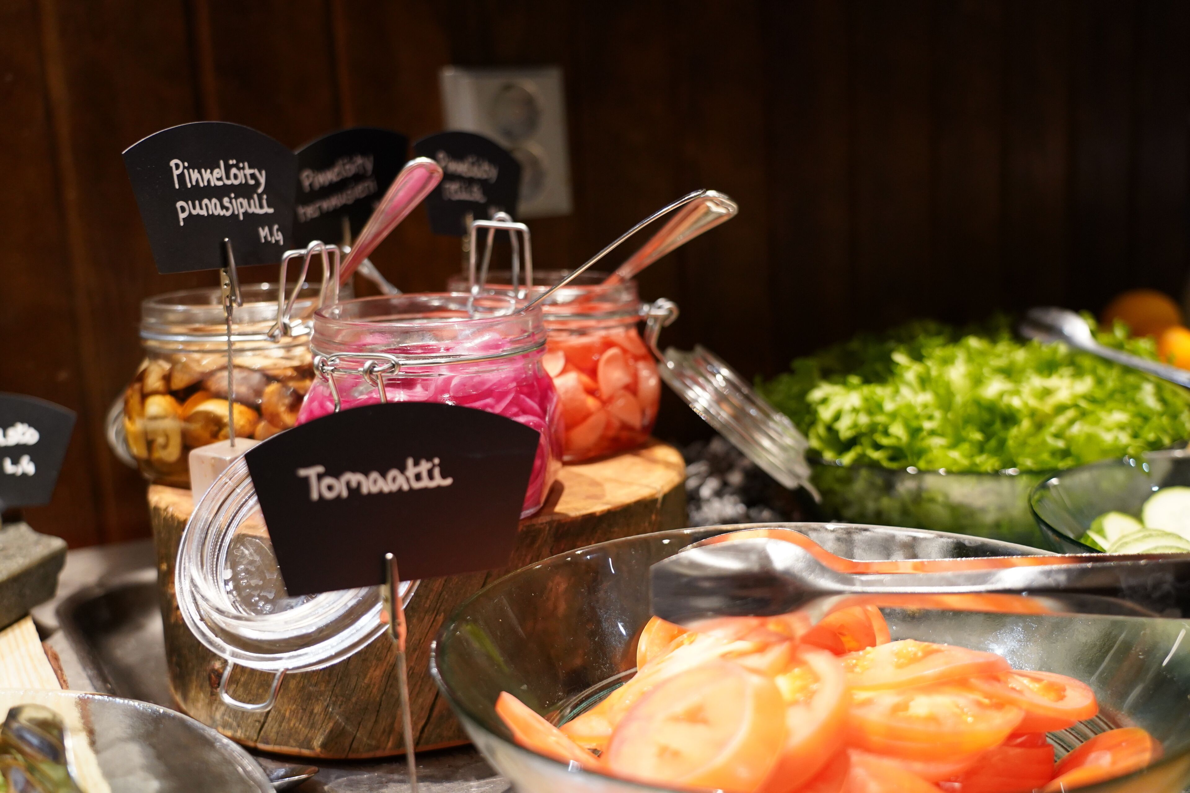A buffet with various food items including jars of pickles, a bowl of sliced tomatoes, and a salad.