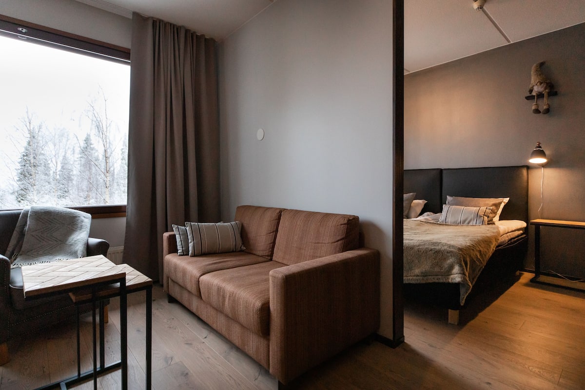 Una habitación moderna con un sofá marrón, una mesita, una cama con cabecero y una ventana con cortinas.