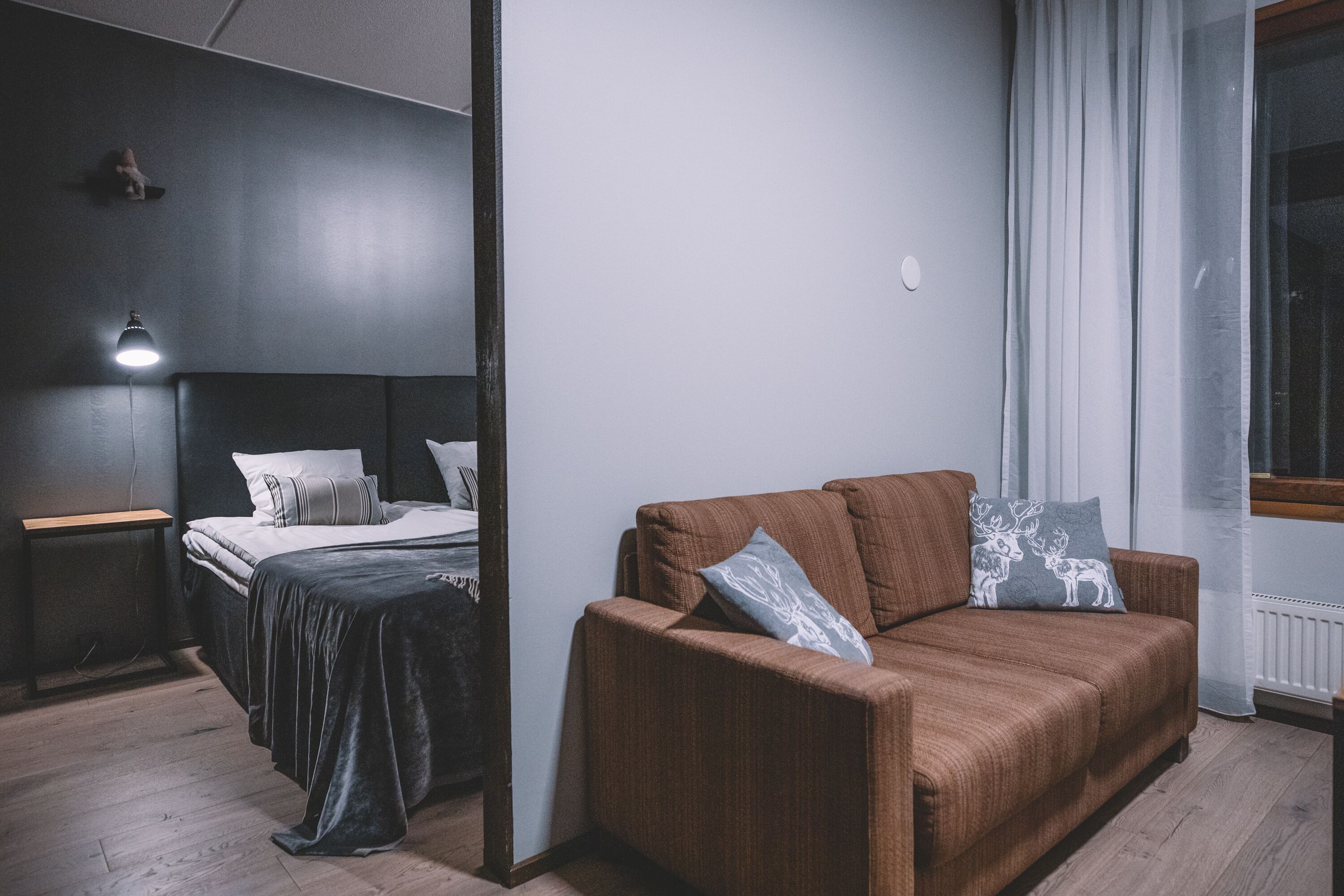 A room with a bed, a brown sofa, a nightstand, a lamp, and a window with curtains.