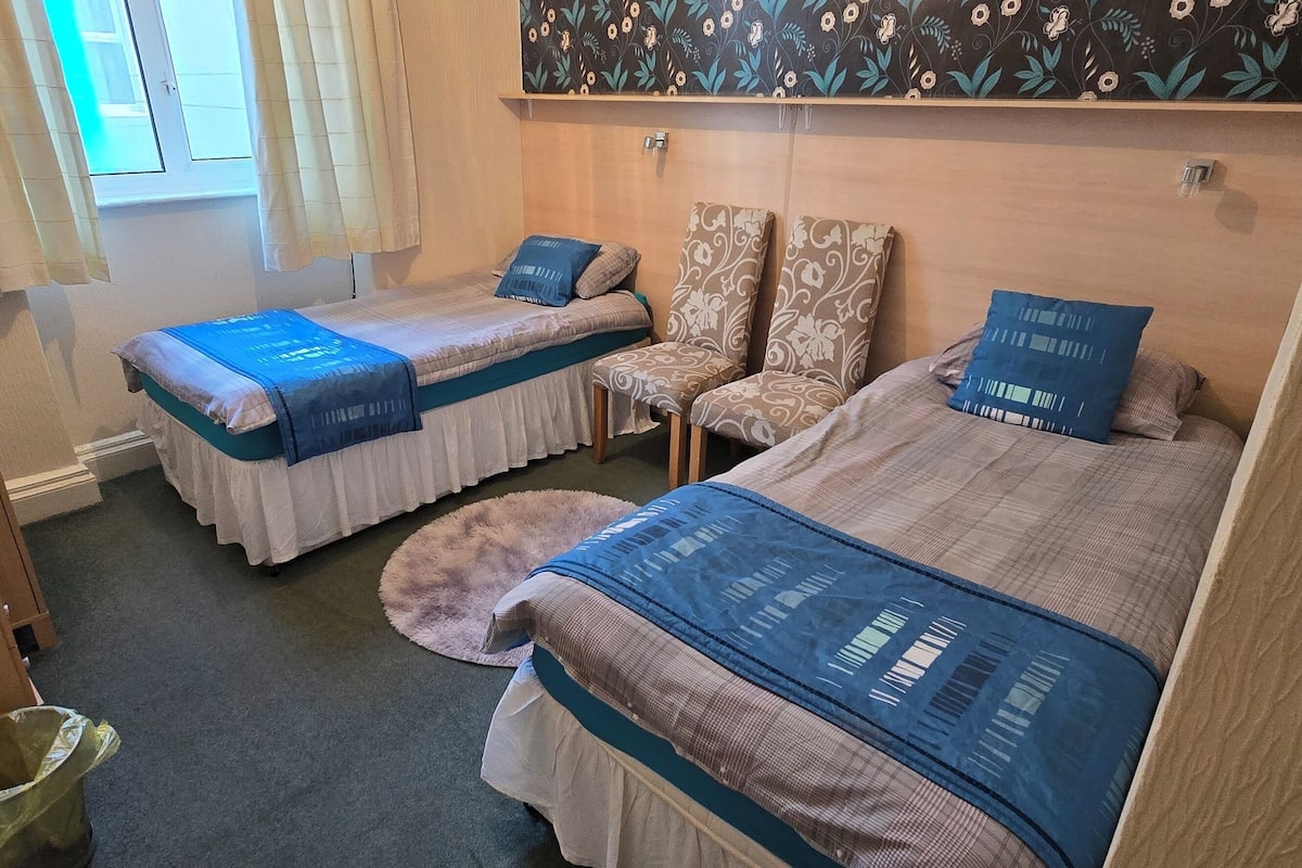 A hotel room with two beds, a small table, a chair, and a trash bin.