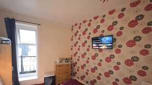 A bedroom with a bed, a wooden dresser, a television mounted on the wall, and a window with curtains. - The Sands Hotel (Blackpool)